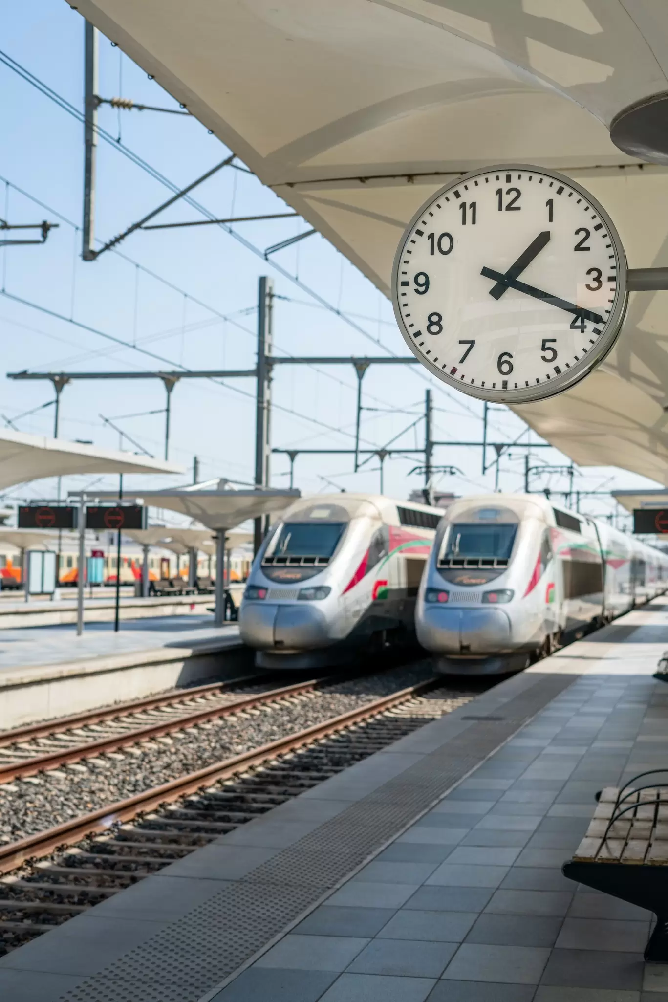 Two silver trains are side by side at an outdoor train station during the daytime. A clock shows 1:20.