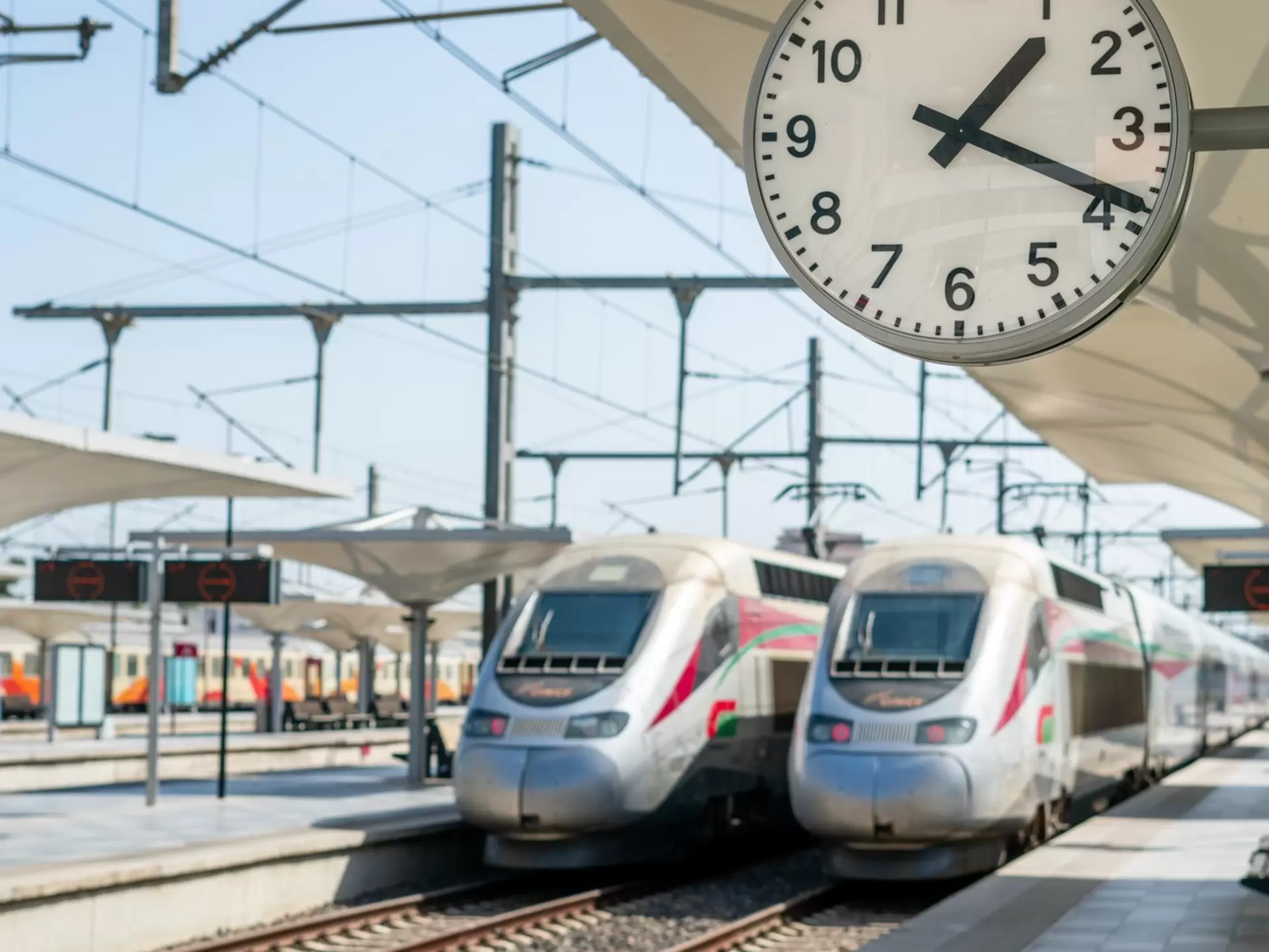 Two silver trains are side by side at an outdoor train station during the daytime. A clock shows 1:20.