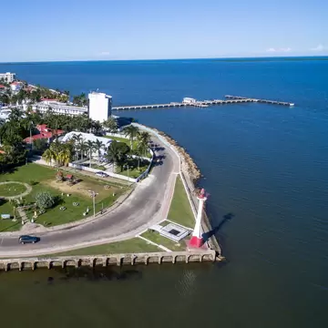 A walkway follows the curve of the coastline, with a red and white lighthouses and palm trees: The waterfront in Belize City. 