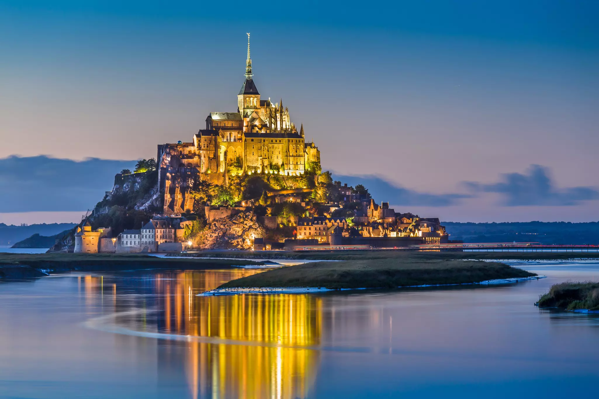 An islet crowned by a monastery stands tall on the sand at low tide in the twilight.