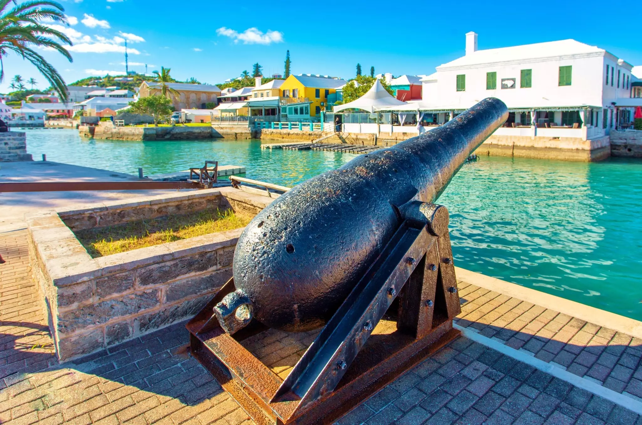 A historic cannon is displayed in the port of a city. Pastel-colored houses are seen across the water.