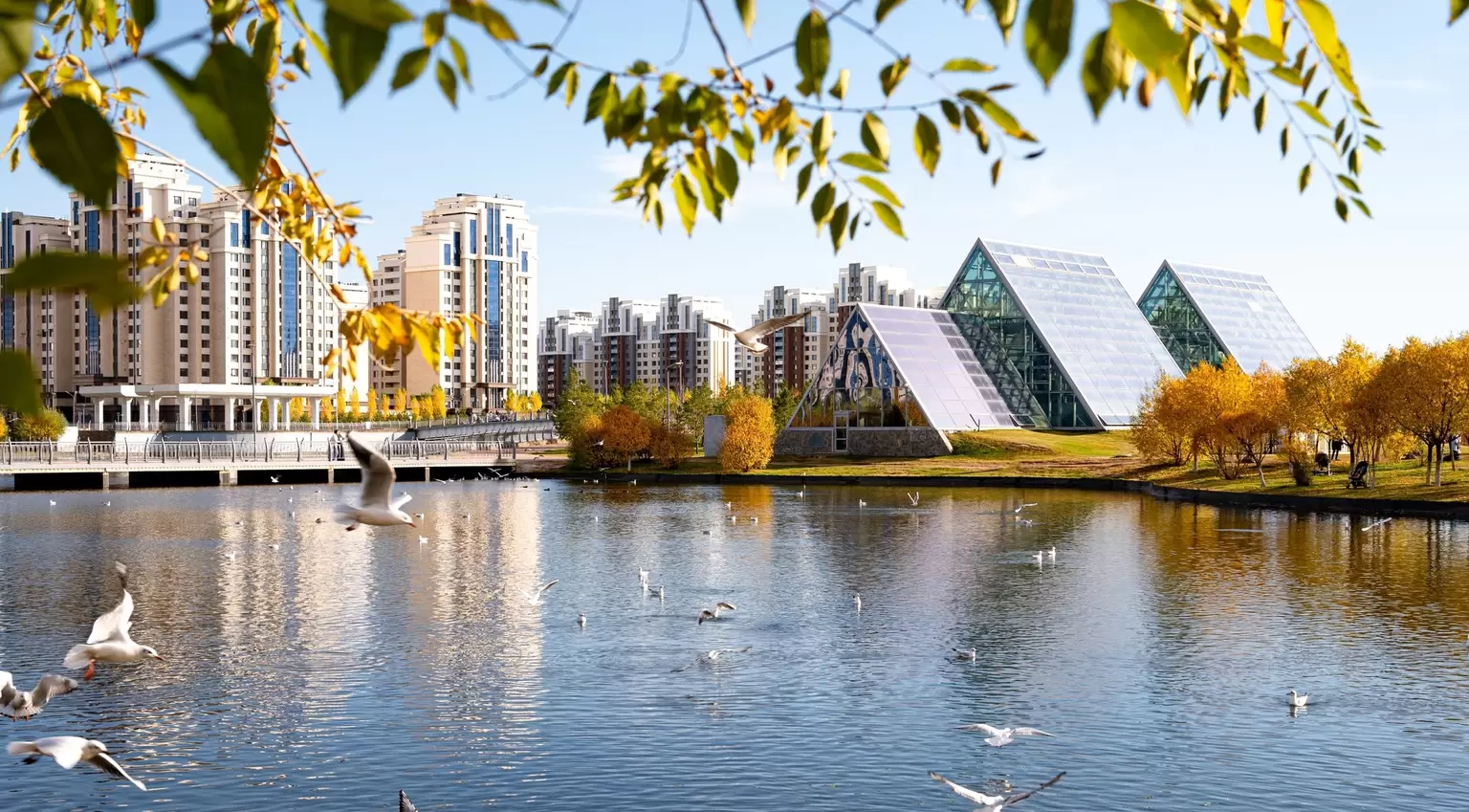 View of the greenhouses with tropical plants in the Astana Botanical Garden across the pond, with flying seagulls on a warm autumn day. Kazakhstan
