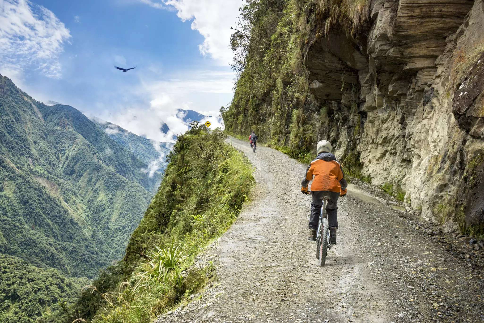 A cyclist follows a single gravel track dug into the side of a mountain as a large bird of prey flies overhead