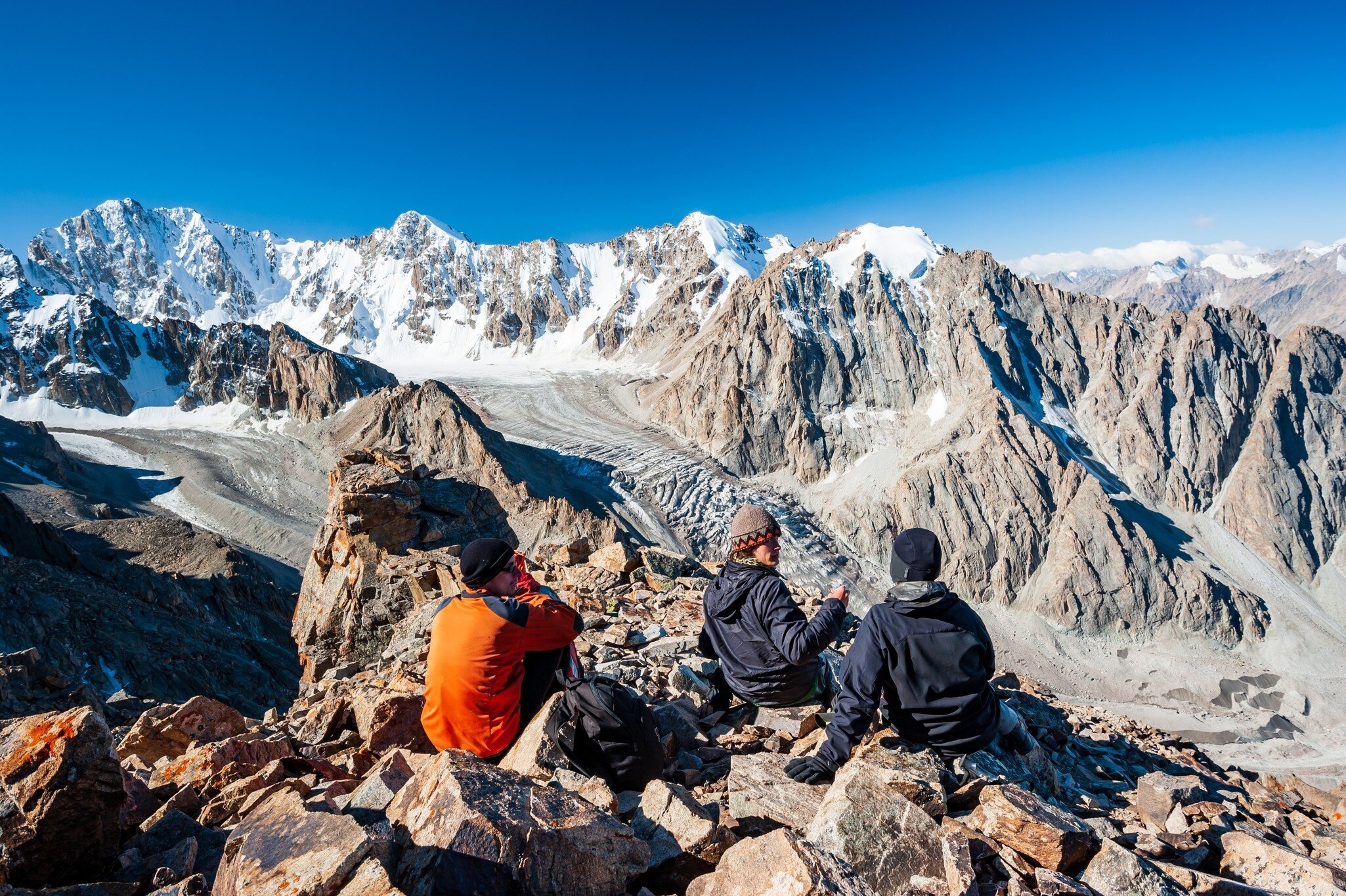 A group of trekkers sit on a rocky outcrop at the top of a mountain overlooking a glacier valley. Spiky peaks, many covered with snow, like beyond the valley.