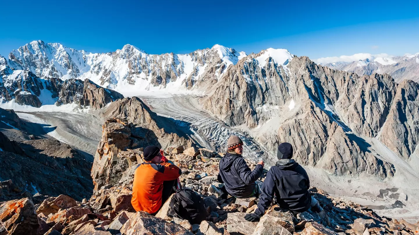 A group of trekkers sit on a rocky outcrop at the top of a mountain overlooking a glacier valley. Spiky peaks, many covered with snow, like beyond the valley.