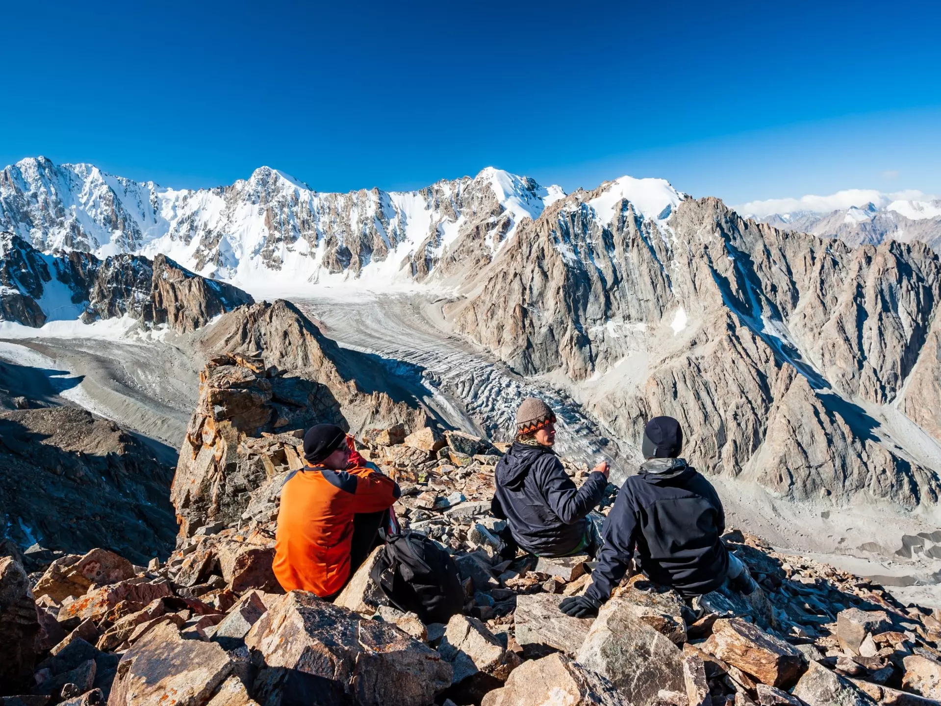 A group of trekkers sit on a rocky outcrop at the top of a mountain overlooking a glacier valley. Spiky peaks, many covered with snow, like beyond the valley.