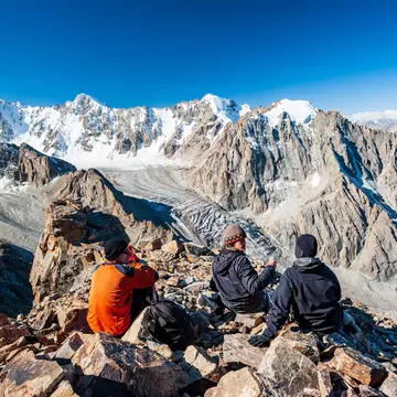 A group of trekkers sit on a rocky outcrop at the top of a mountain overlooking a glacier valley. Spiky peaks, many covered with snow, like beyond the valley.
