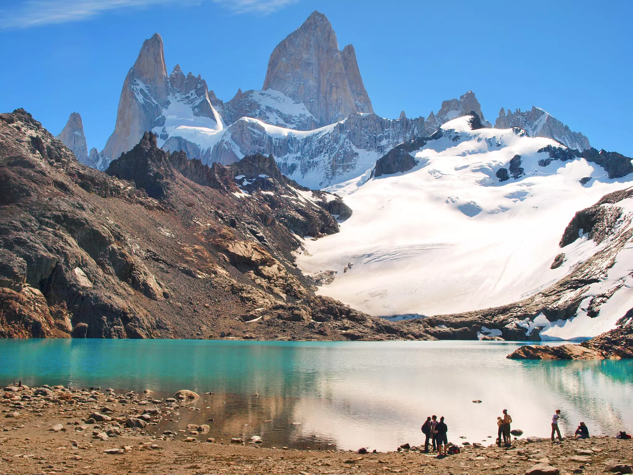 A group of people stand on rocky ground by a turquoise lake facing snow-covered peaks.