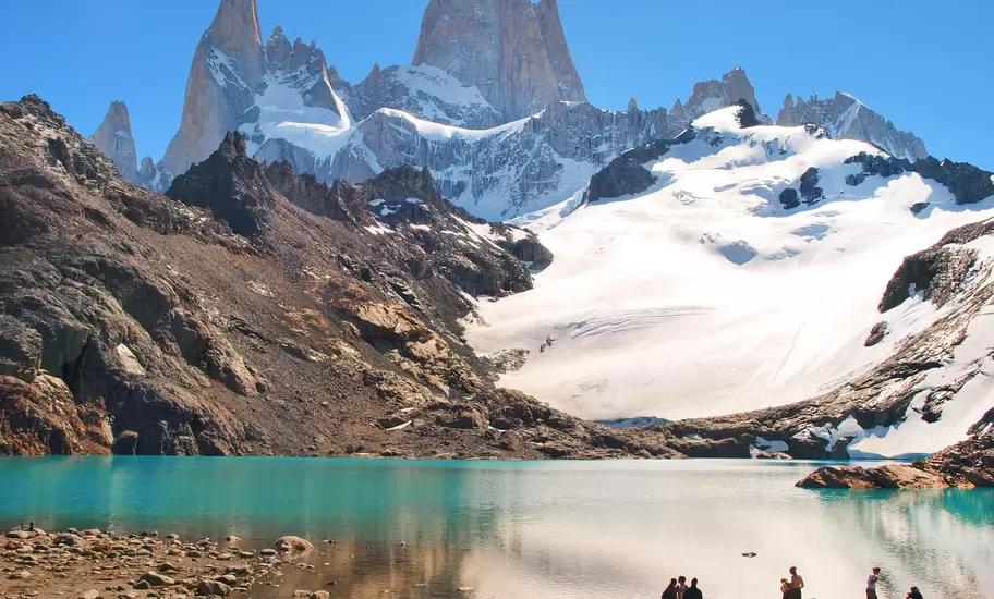 A group of people stand on rocky ground by a turquoise lake facing snow-covered peaks.