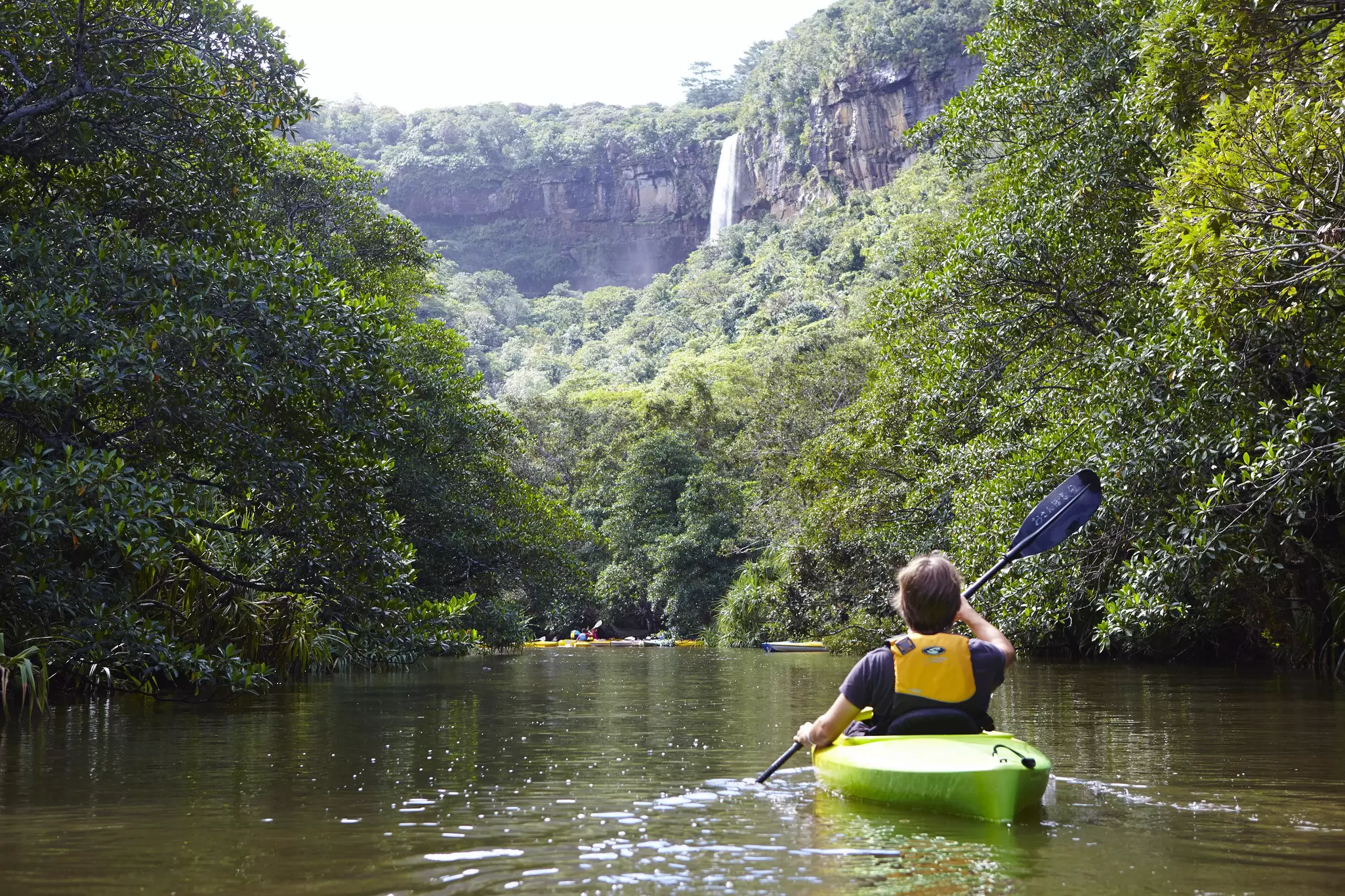 A kayaker paddles a green boat on a river in a lush forest.