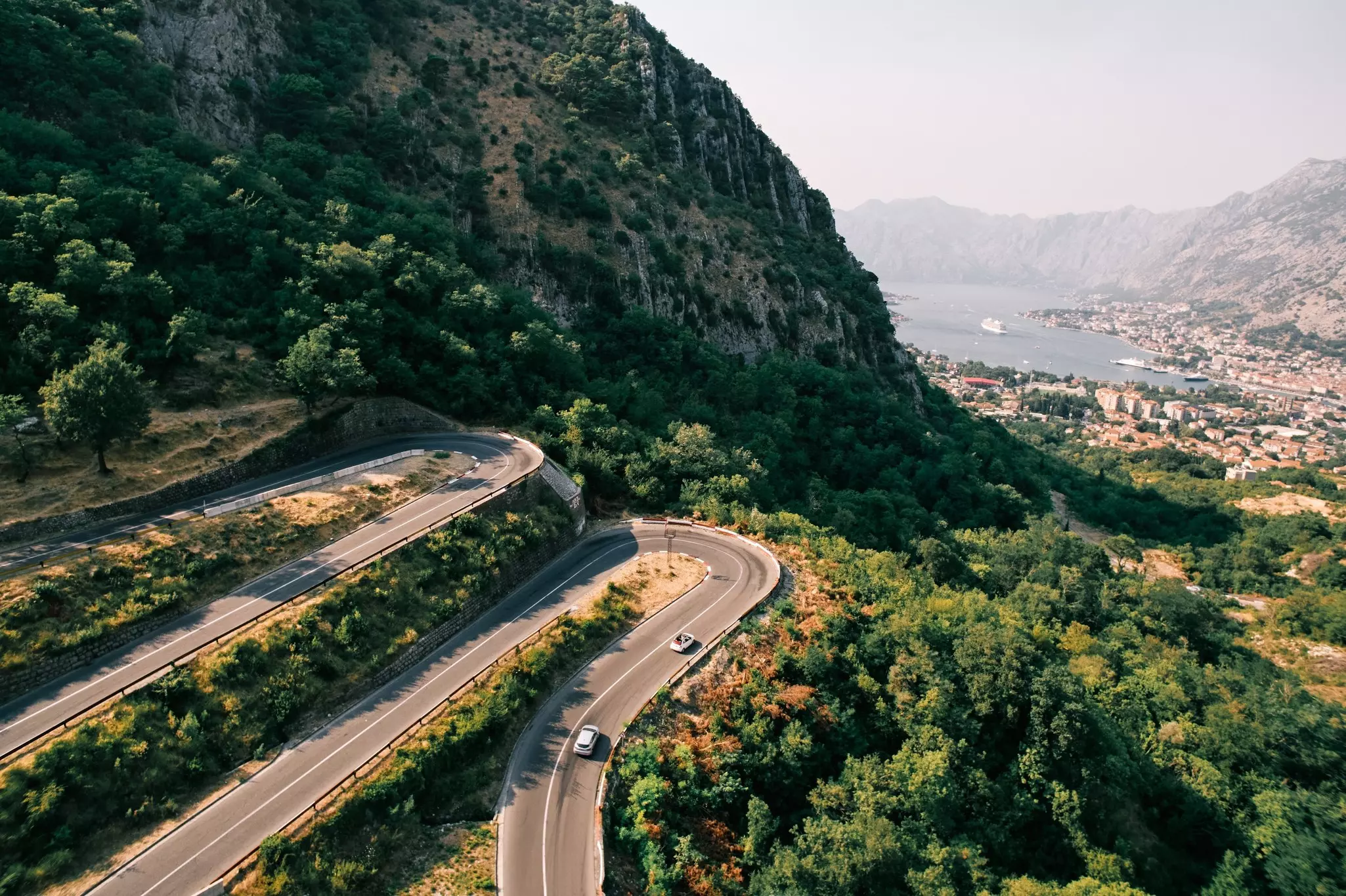 Two cars on a road with sharp switchbacks up a green mountain, with a town in the distance by the shore.