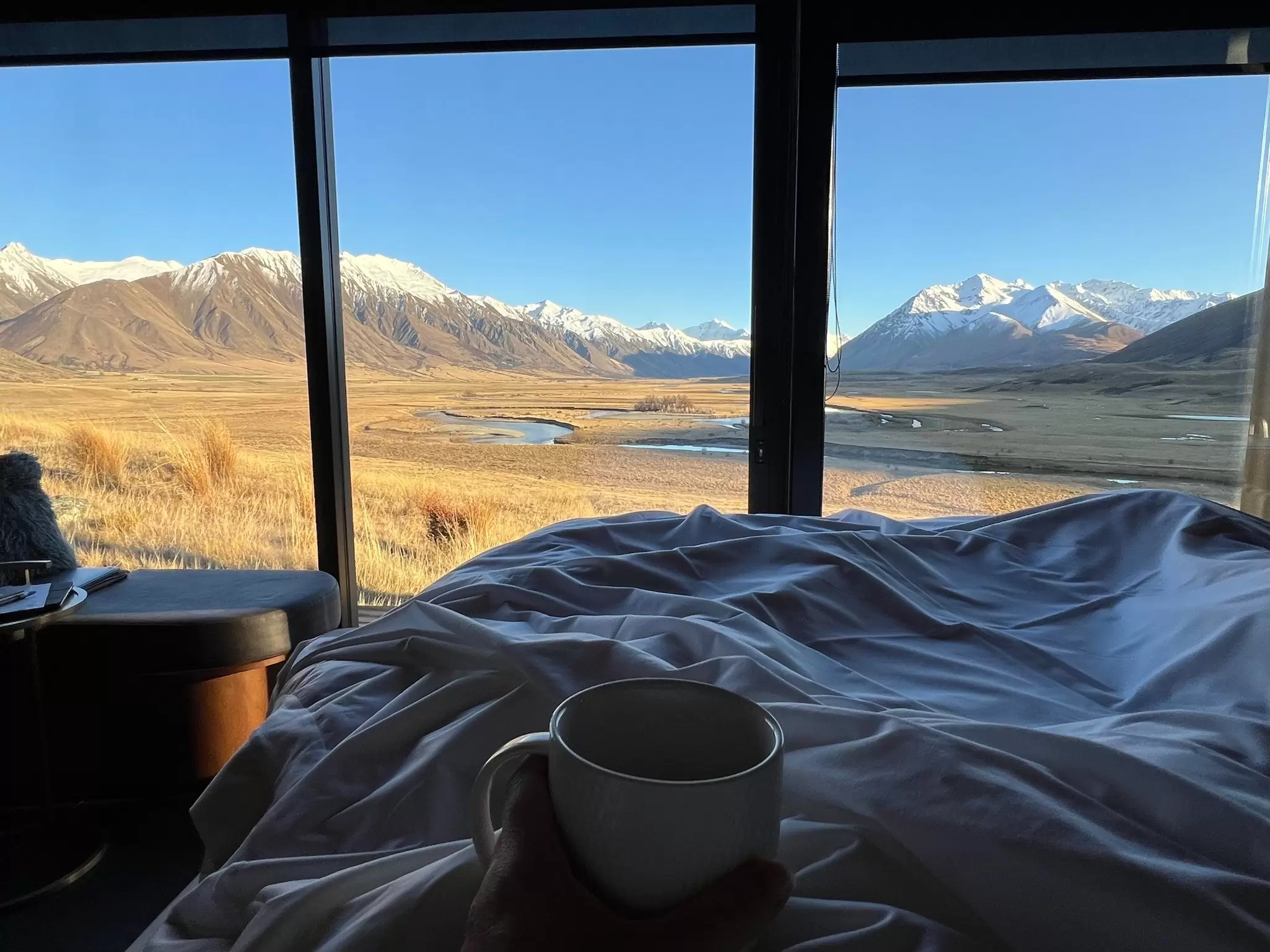 A point of view of a person holding a coffee cup in bed looking out at a river and mountains, Lindis luxury lodge in the Ahuriri Valley, New Zealand