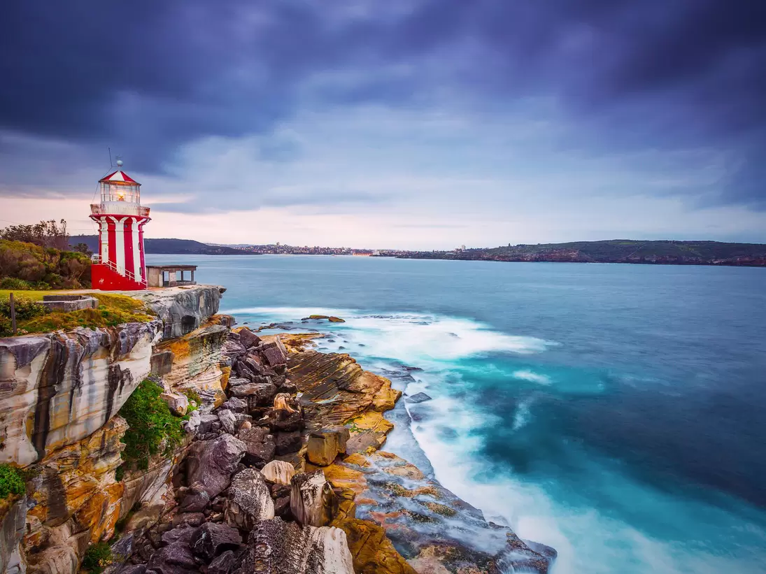 A red-and-white striped lighthouse on a sandstone headland above breaking surf under storm clouds.