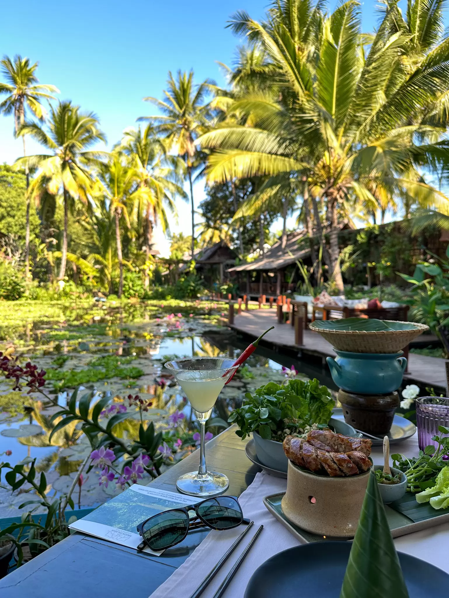 A cocktail on a table with a banana leaf and plates of food beside a lily pond surrounded by tropical vegetation in the sunshine.