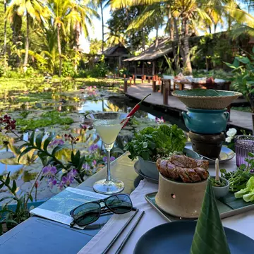 A cocktail on a table with a banana leaf and plates of food beside a lily pond surrounded by tropical vegetation in the sunshine.