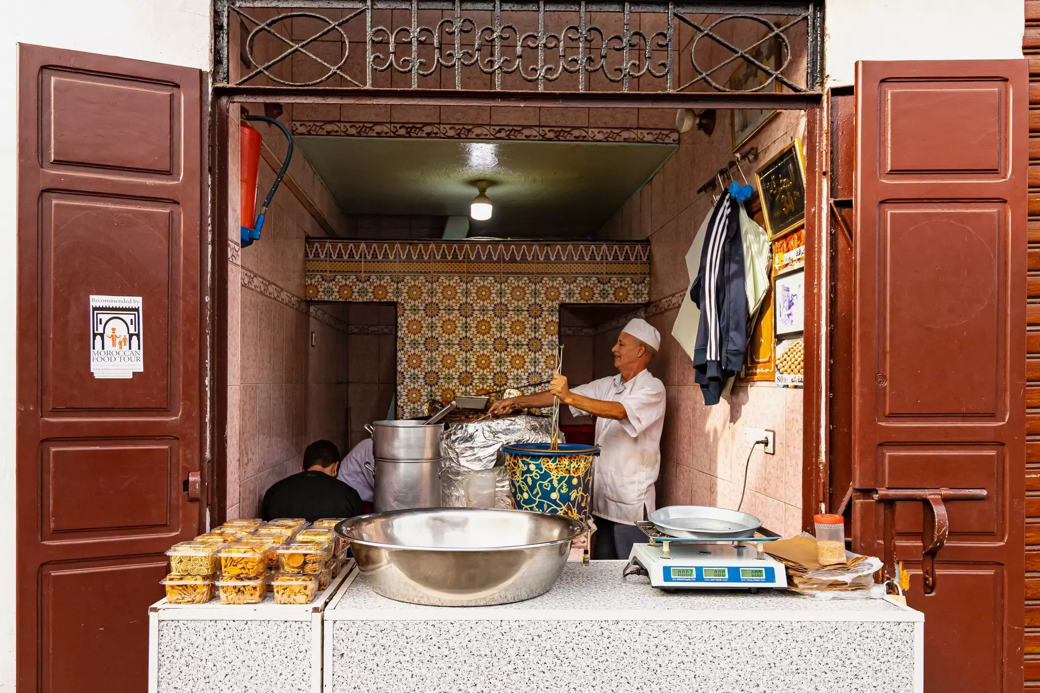 A worker at a large pot set inside a large open doors; another large metal bowl is on a table at the street.