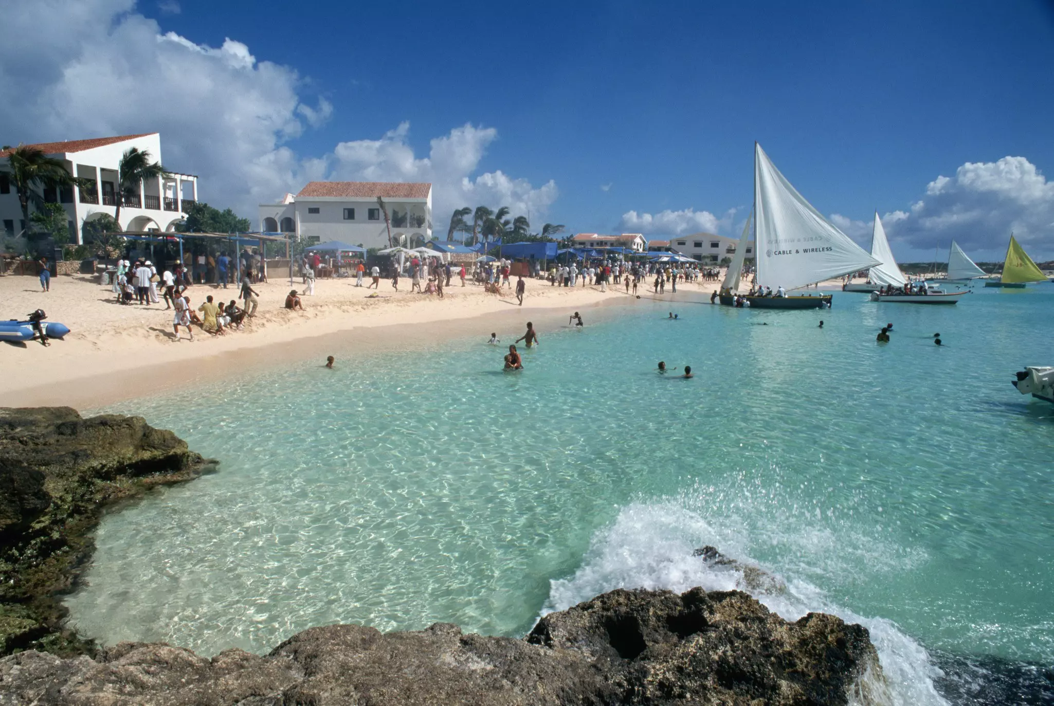 Small boats with large sails docked near the shallow water of a golden-sand beach as many people gather on the shore.