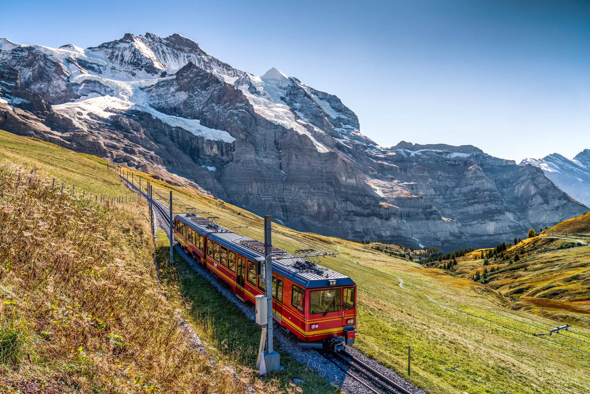 A red train running across a green hillside in Switzerland; rocky peaks with patches of snow are in the background.