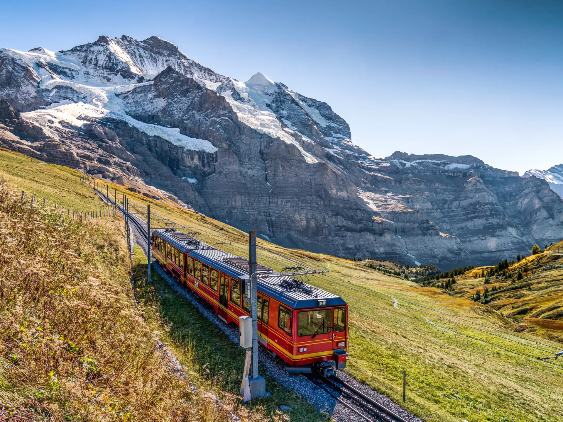A small red train follows a track on a mountain slope, with views of snow-covered peaks across a valley.
