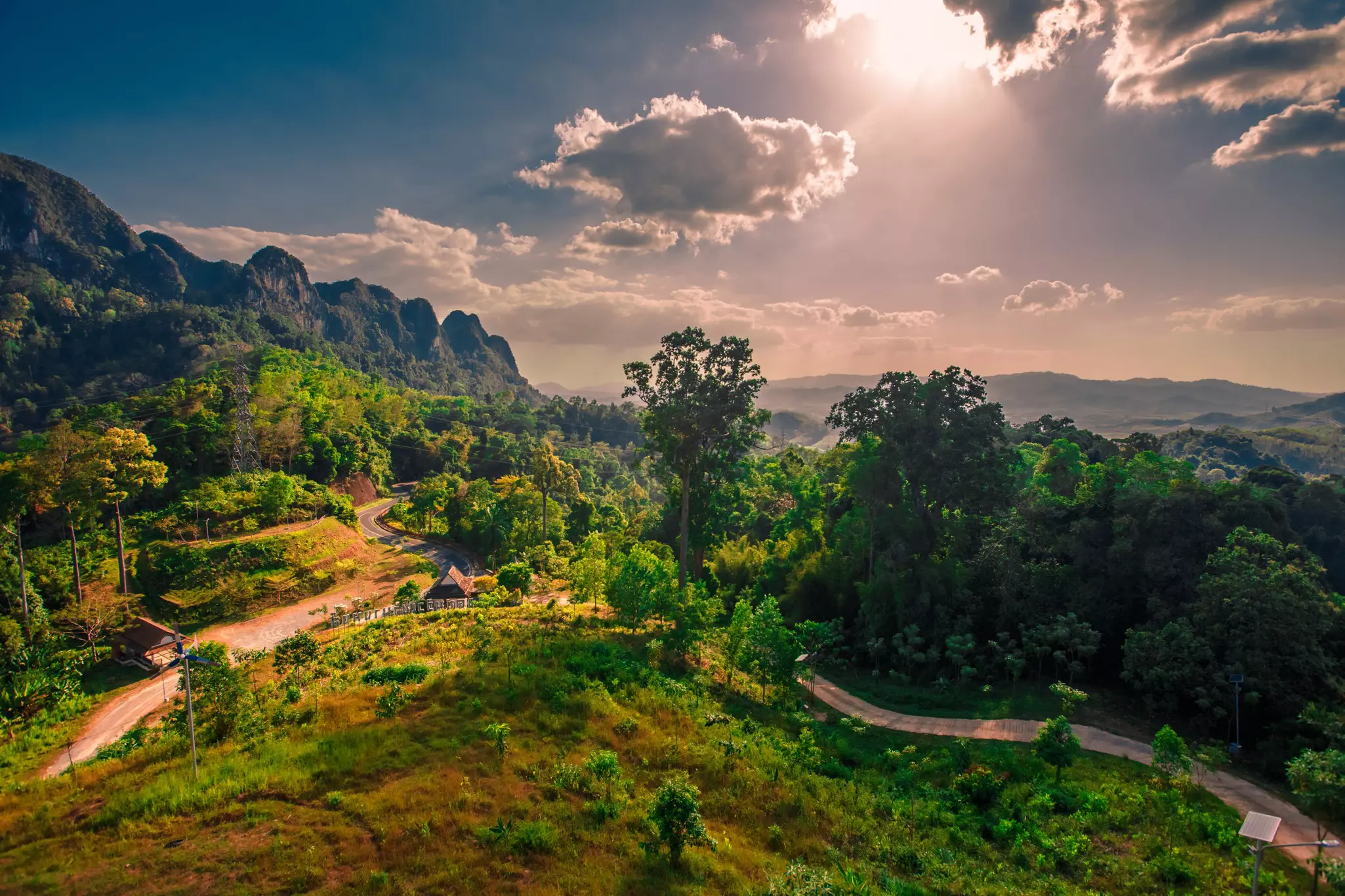 Sunrise in a lush, mountainous landscape in Thailand; a road snakes diagonally across between trees and a grassy hillside.