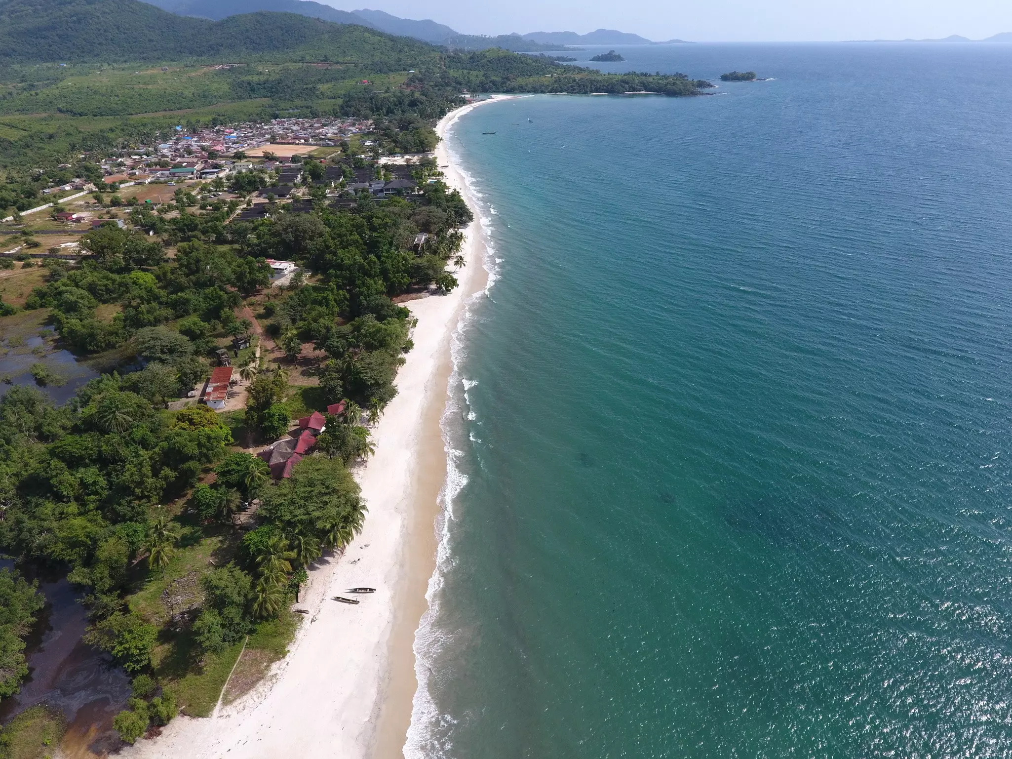 An aerial view of a long strip of beach, with the ocean seen to the right and lush vegetation to the left.