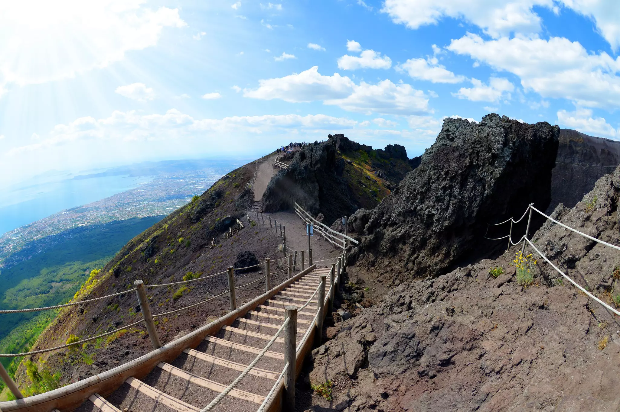 View of a Mount Vesuvius hiking trail featuring a wooden and dirt stairway and handrails with a bay and clouds in the distance.