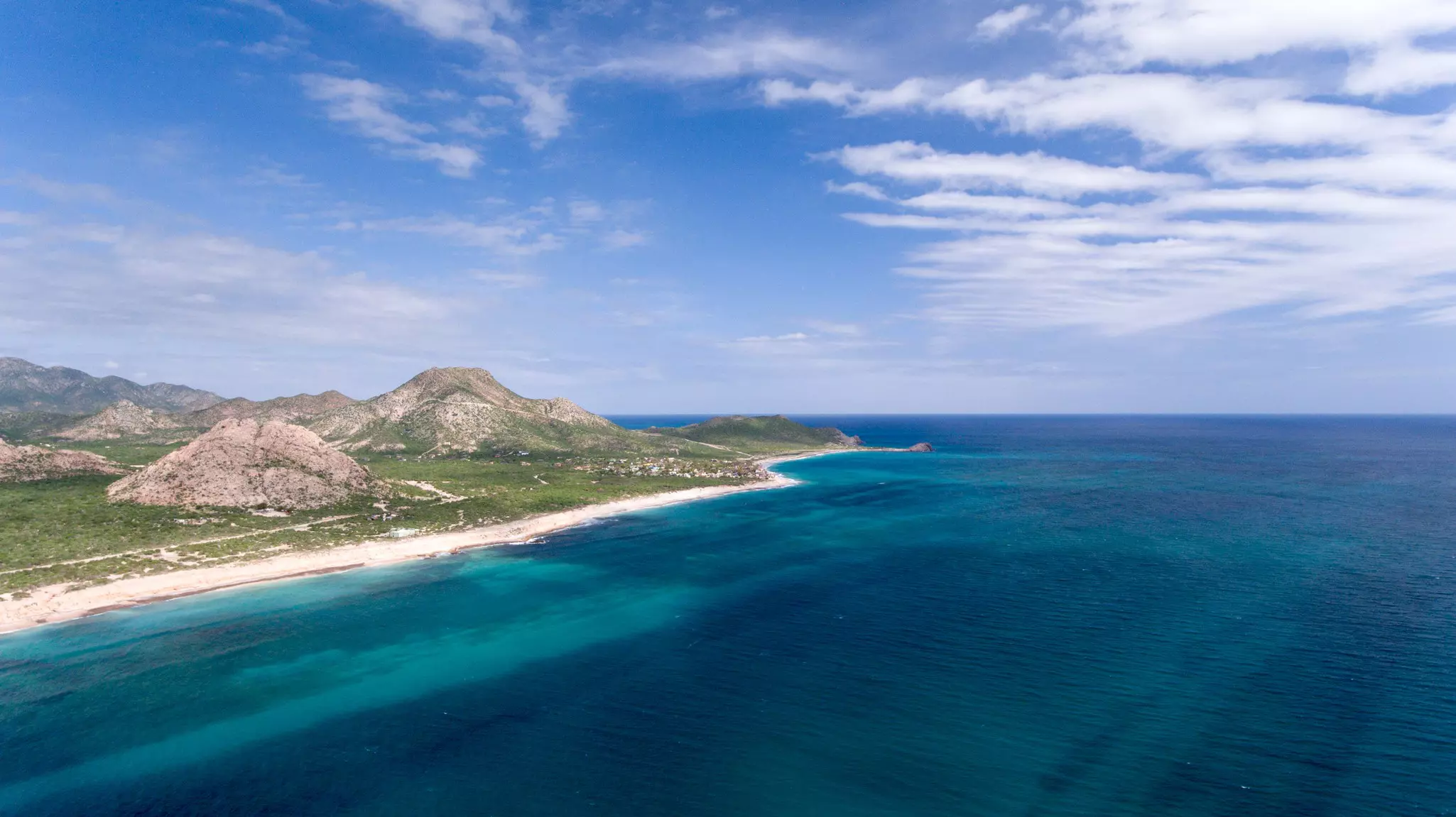 Aerial view of dark blue ocean with narrow sandy beach, grassland and rocky hills to the left on a sunny day.