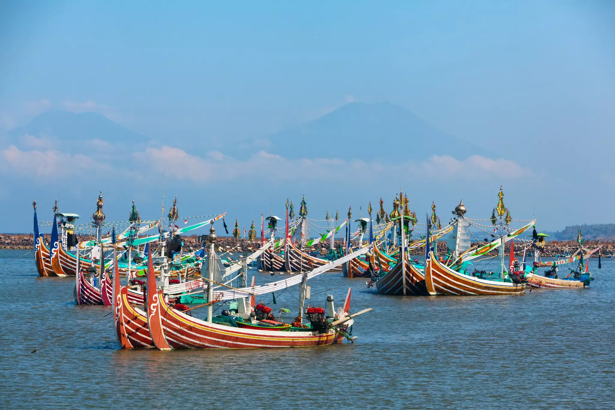 Brightly colored fishing boats are moored off a tropical island. A huge volcano is seen in the haze in the distance.