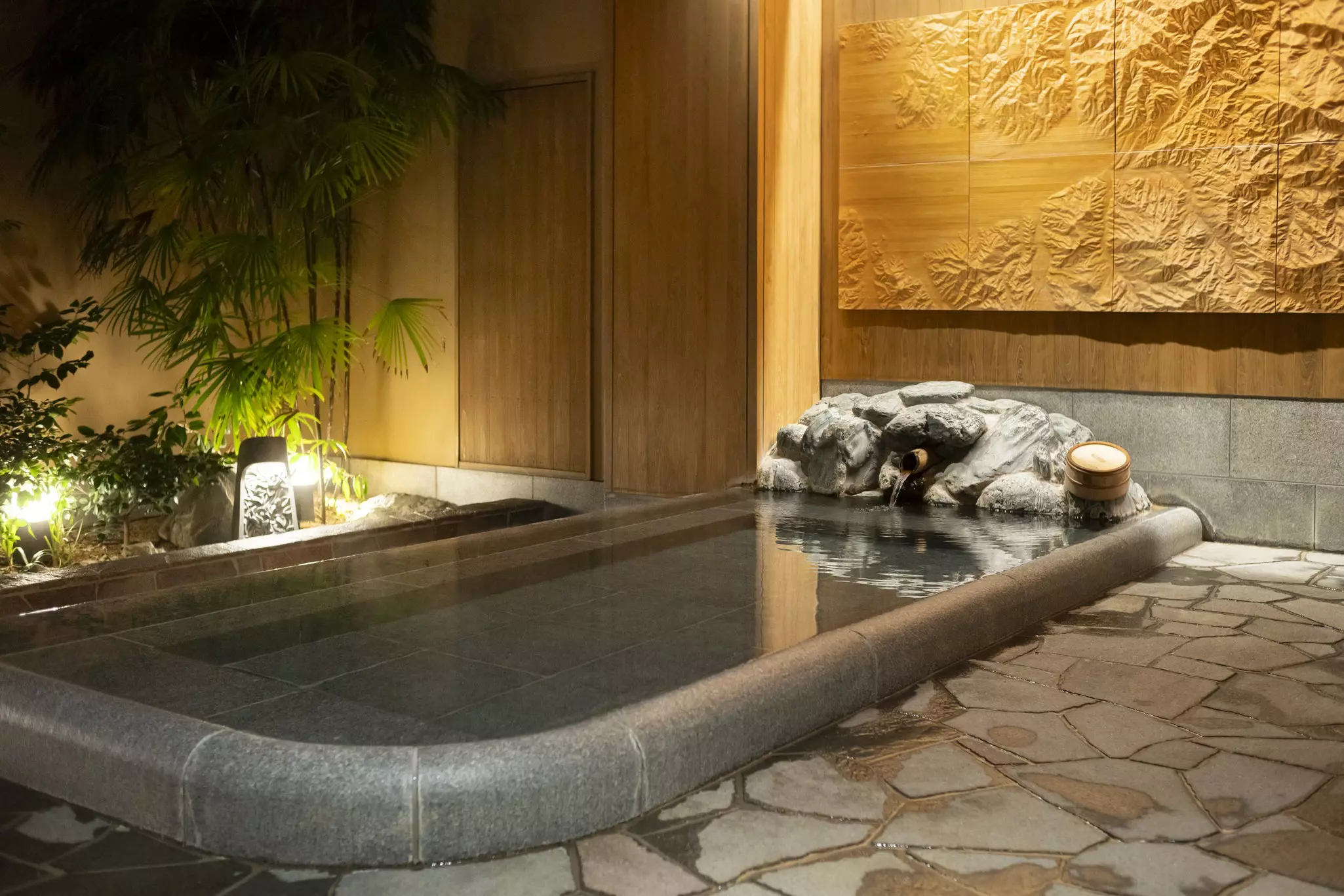 Large gray stone indoor bath surrounded by flat stones with palms and other plants in the distance and wooden relief artwork on the wall.