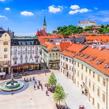 An old-town square with a stone fountain and pavement cafés, ringed by pastel facades and red roofs, with a white castle on the hill behind.