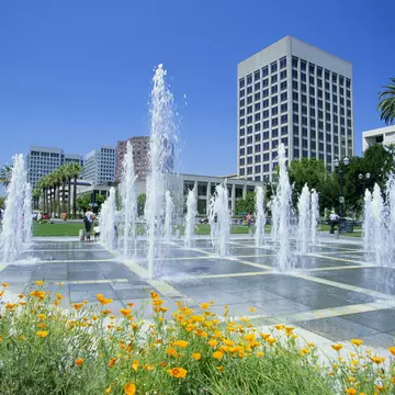 Multiple water spouts rising up from flat sidewalk fountain on a sunny day with tall city buildings in the distance.