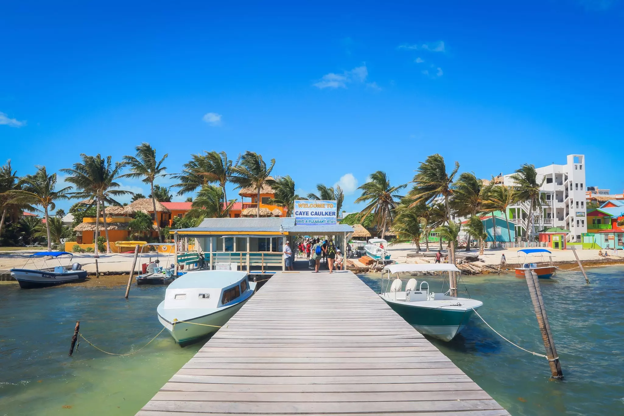 People walk down a long dock with boats tied on either side toward a structure by a sandy beach.