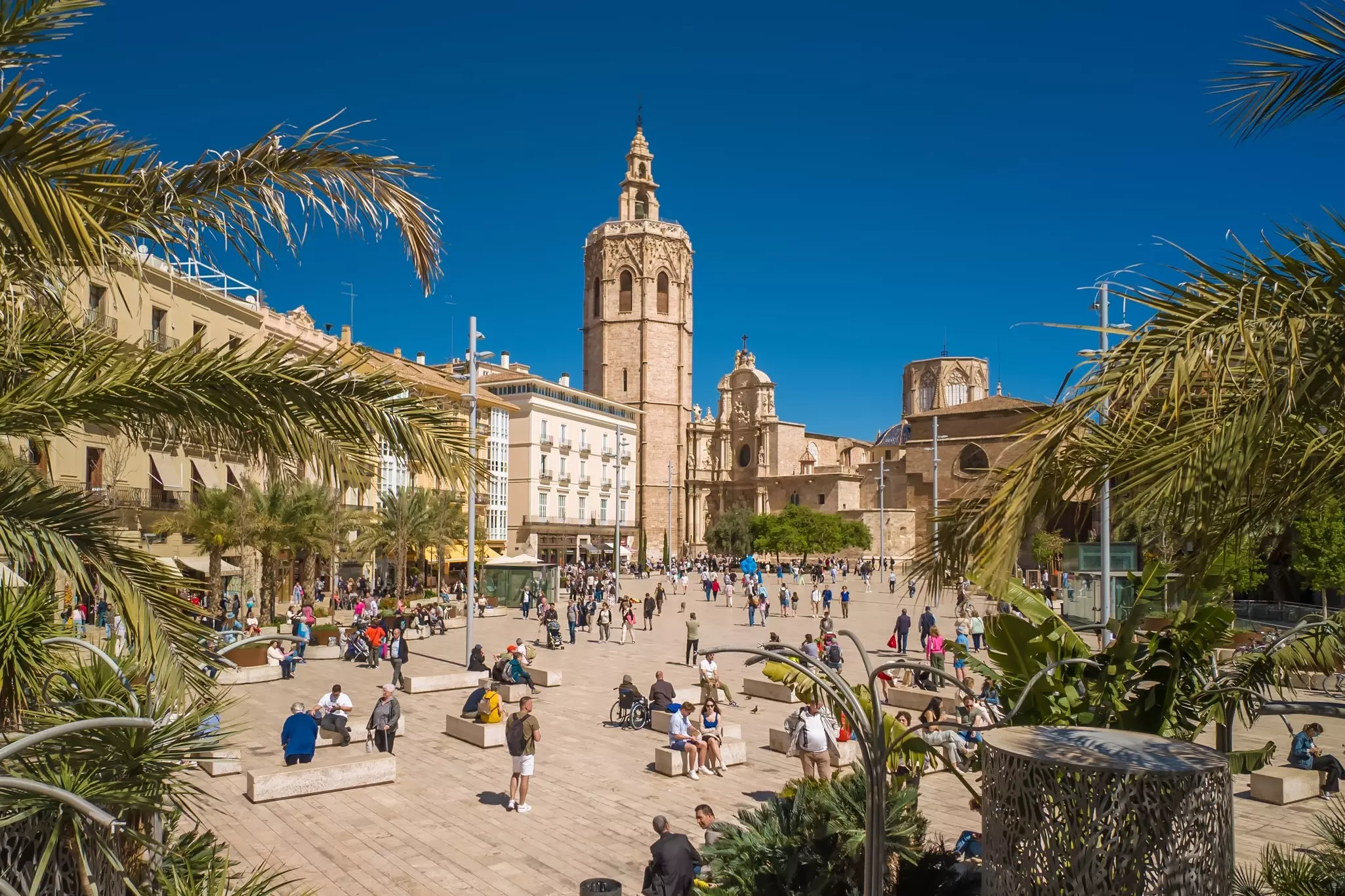 People gather in a plaza overlooked by a cathedral bell tower on a sunny day