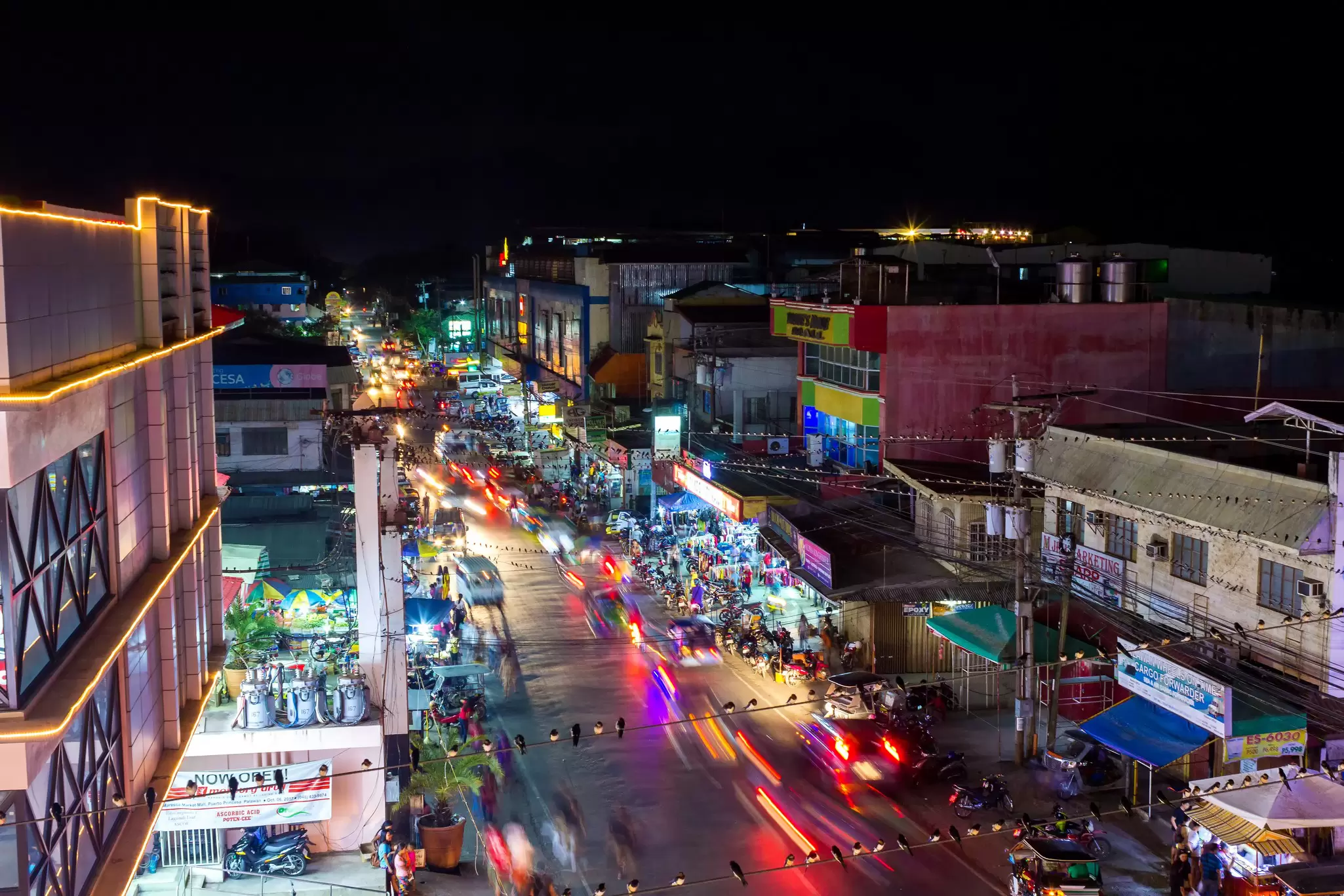 Aerial view of a busy city street at night. The street is lined with low-profile buildings. Rear lights of cars are blurry in motion.