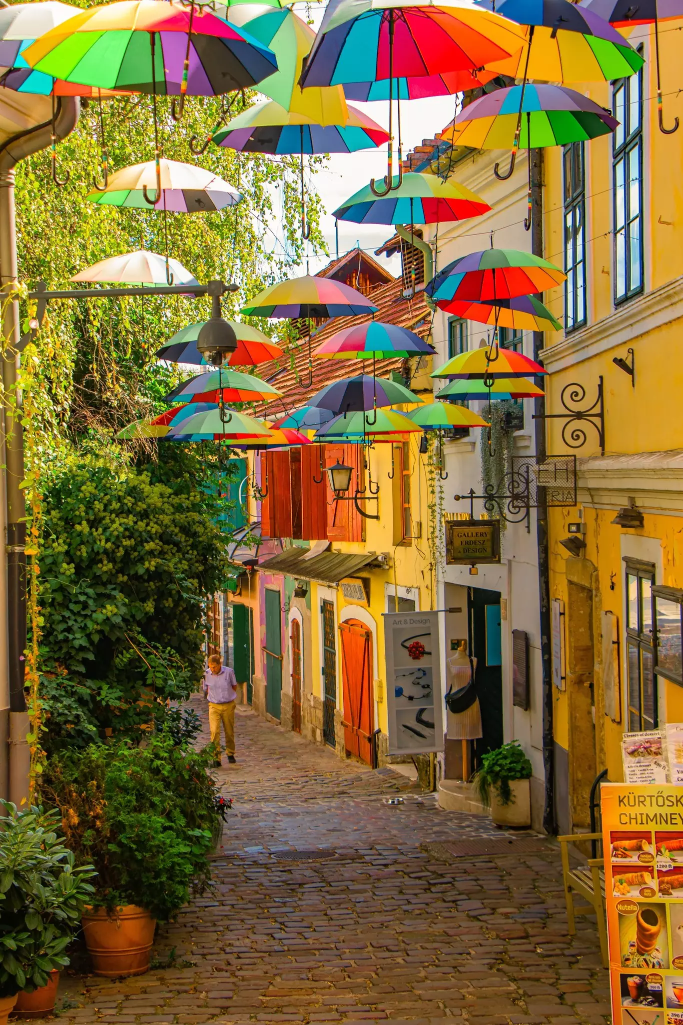 Multicolored umbrellas hang over a cobblestone street with colorful buildings and a single pedestrian.