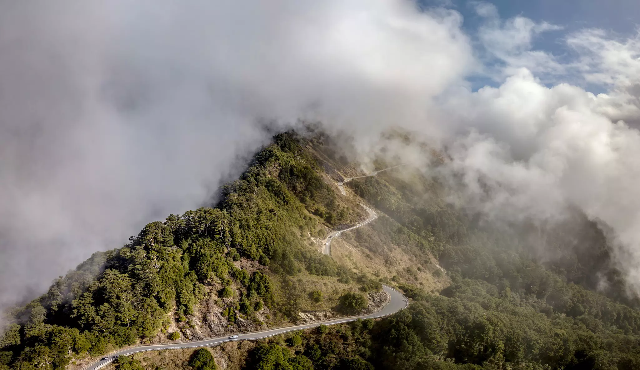 A road winds up the ridge of a tree-covered mountain, with clouds covering higher elevations.