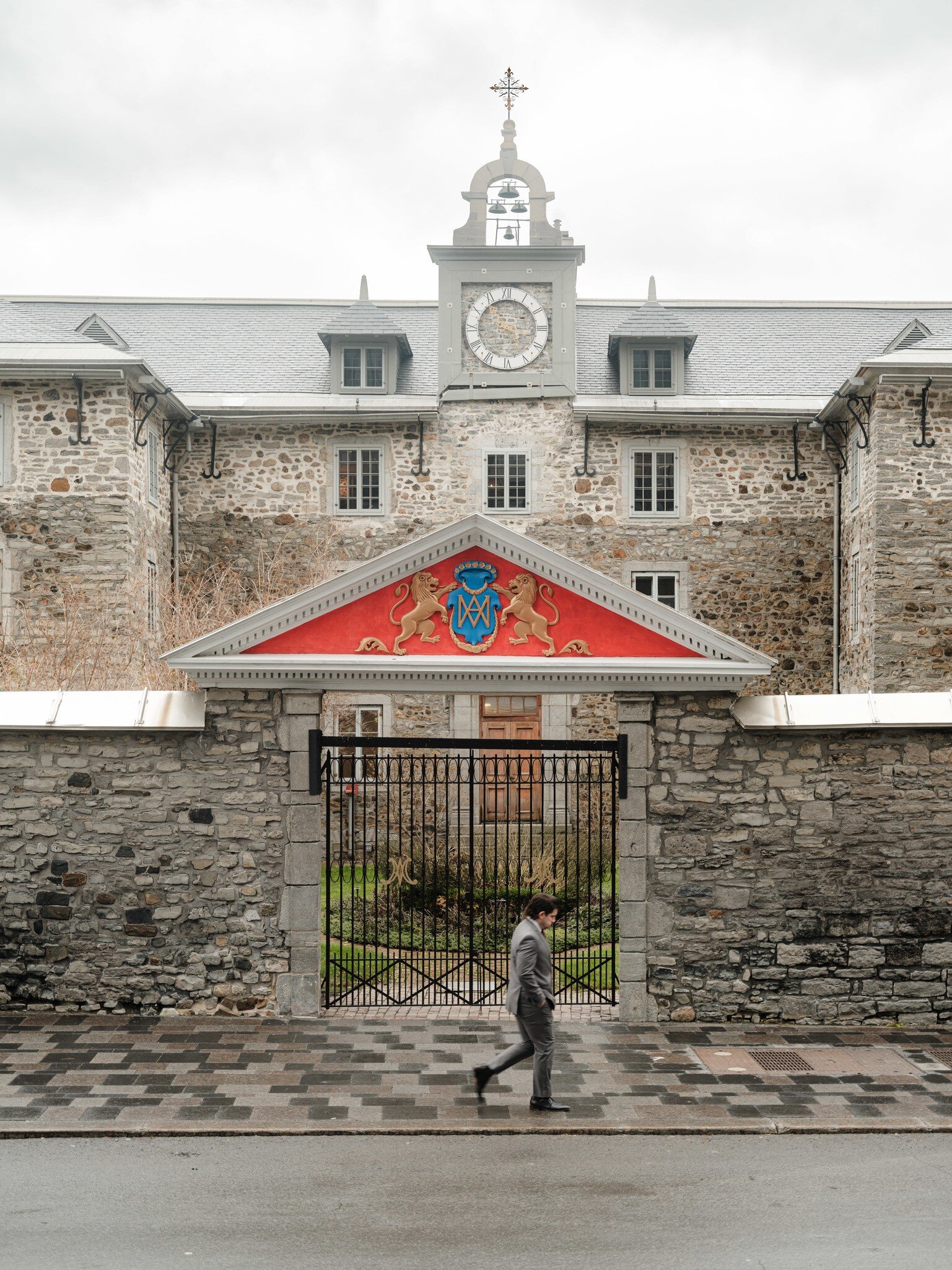 Pedestrian passing an iron gate under a red coat-of-arms pediment in front of a grey stone building with a clock tower.