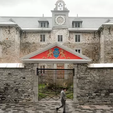Pedestrian passing an iron gate under a red coat-of-arms pediment in front of a grey stone building with a clock tower.