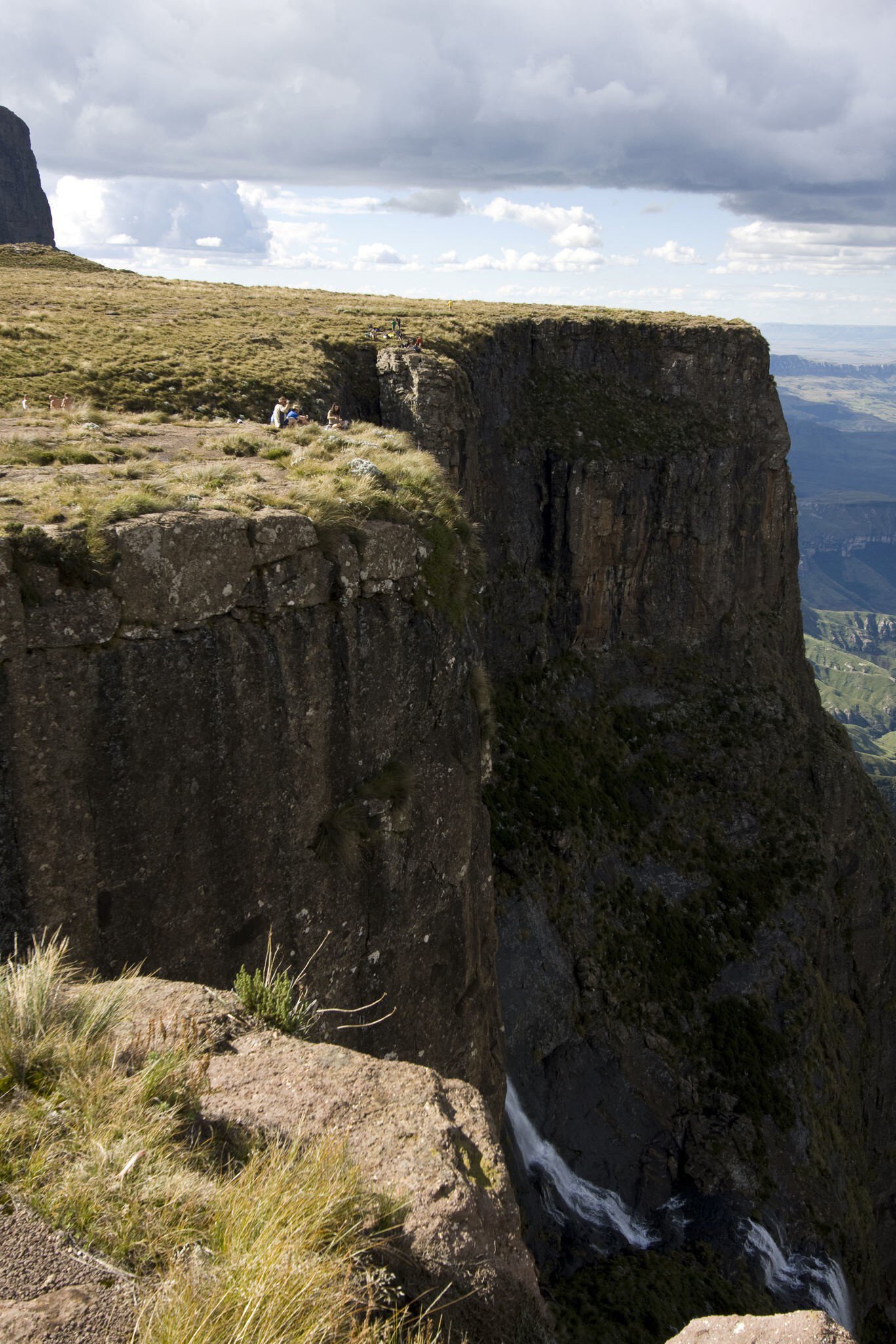 Hikers sit on a grassy clifftop above a sheer rock wall and a waterfall in the Drakensberg mountains, South Africa.