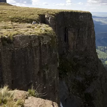 Hikers sit on a grassy clifftop above a sheer rock wall and a waterfall in the Drakensberg mountains, South Africa.