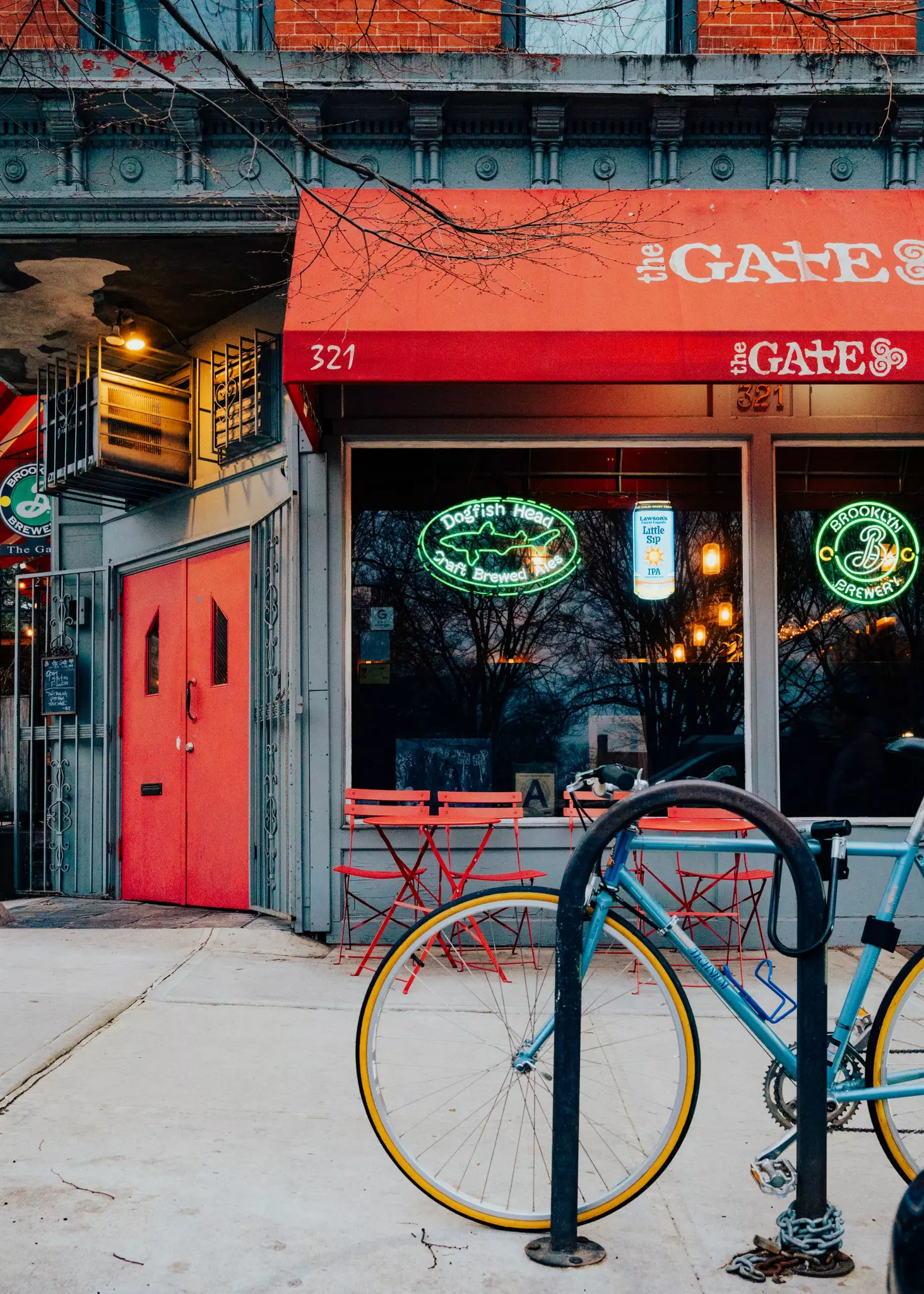 A blue bike is locked to a bike rack in front of a grey buiding with a red door and red chairs. The red awning reads The Gate.
