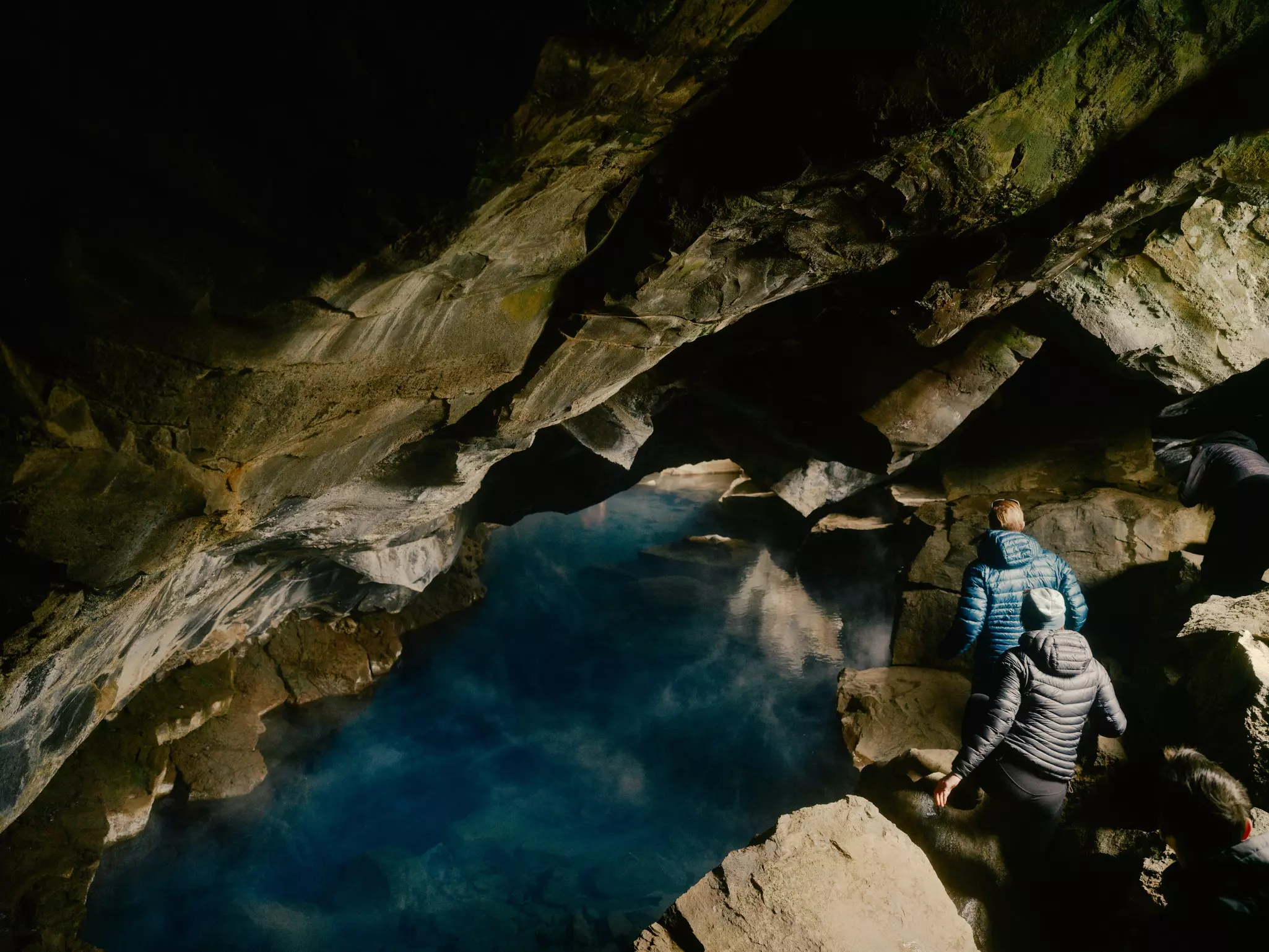 People in winter gear walking in an underground cave with blue water to the left.