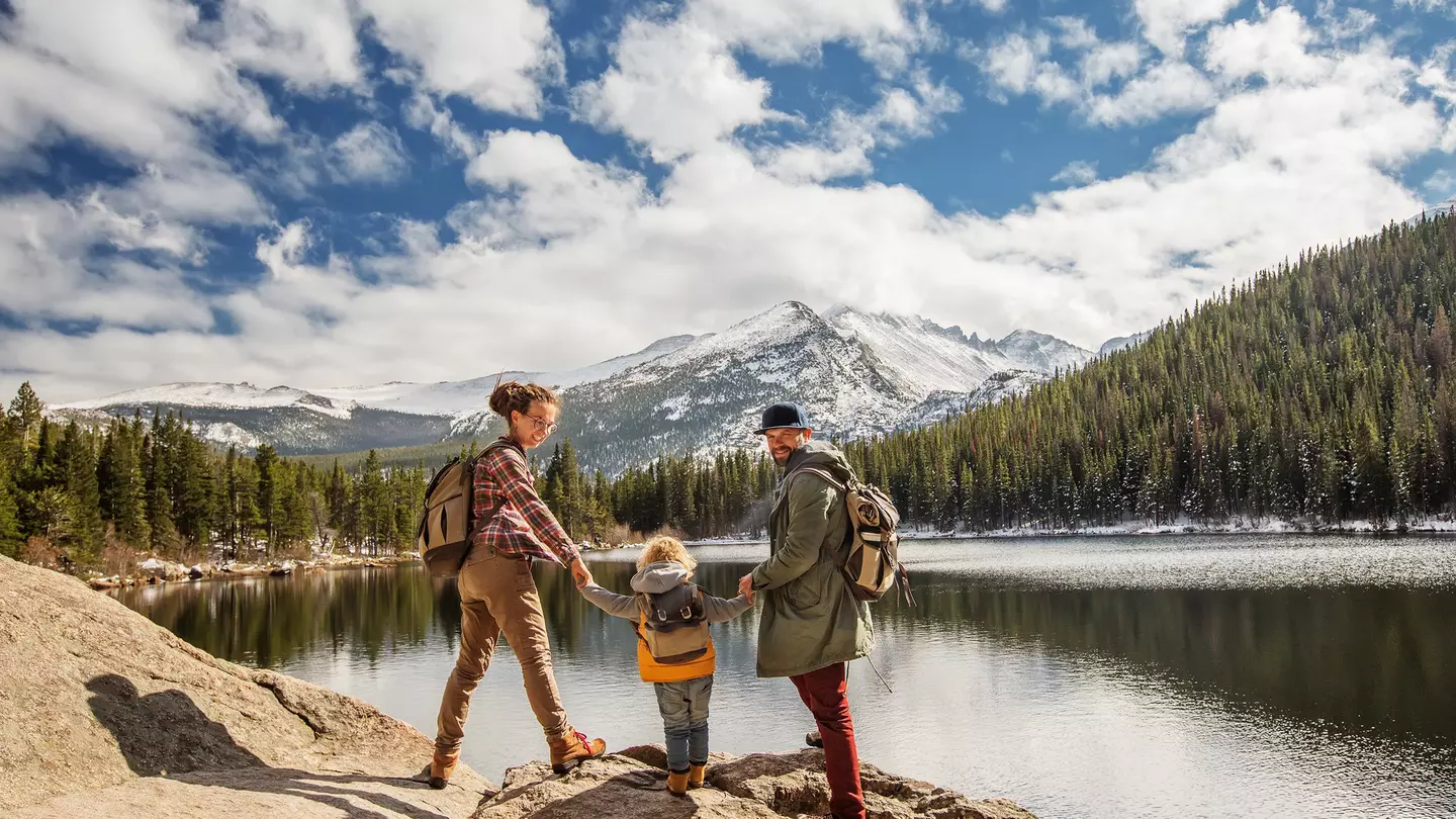 Two parents and a child hike by lake with pines and snowy mountains in the distance at Happy family in Rocky Mountain National Park, Colorado, USA