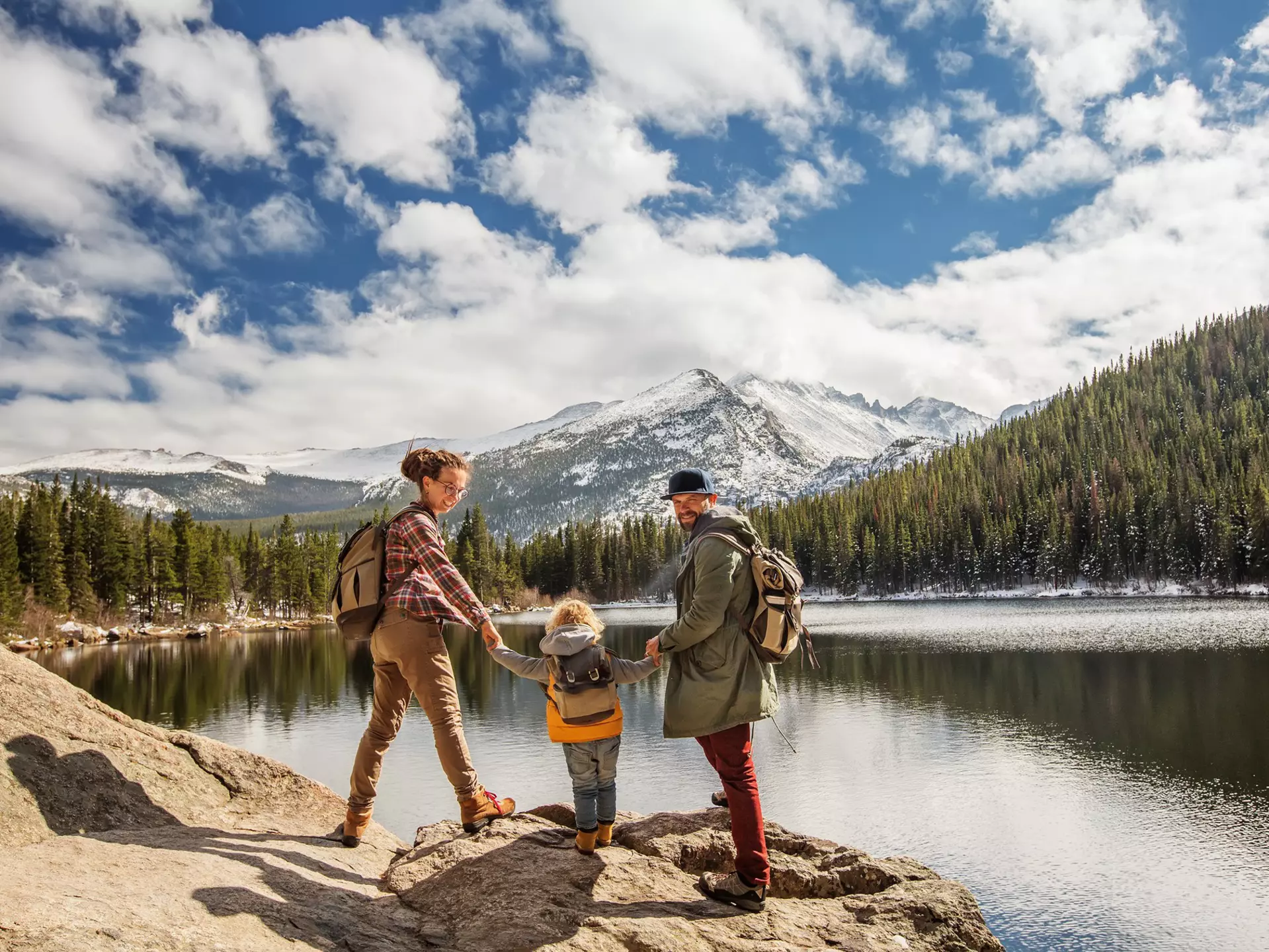 Two parents and a child hike by lake with pines and snowy mountains in the distance at Happy family in Rocky Mountain National Park, Colorado, USA