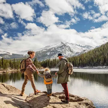 Two parents and a child hike by lake with pines and snowy mountains in the distance at Happy family in Rocky Mountain National Park, Colorado, USA