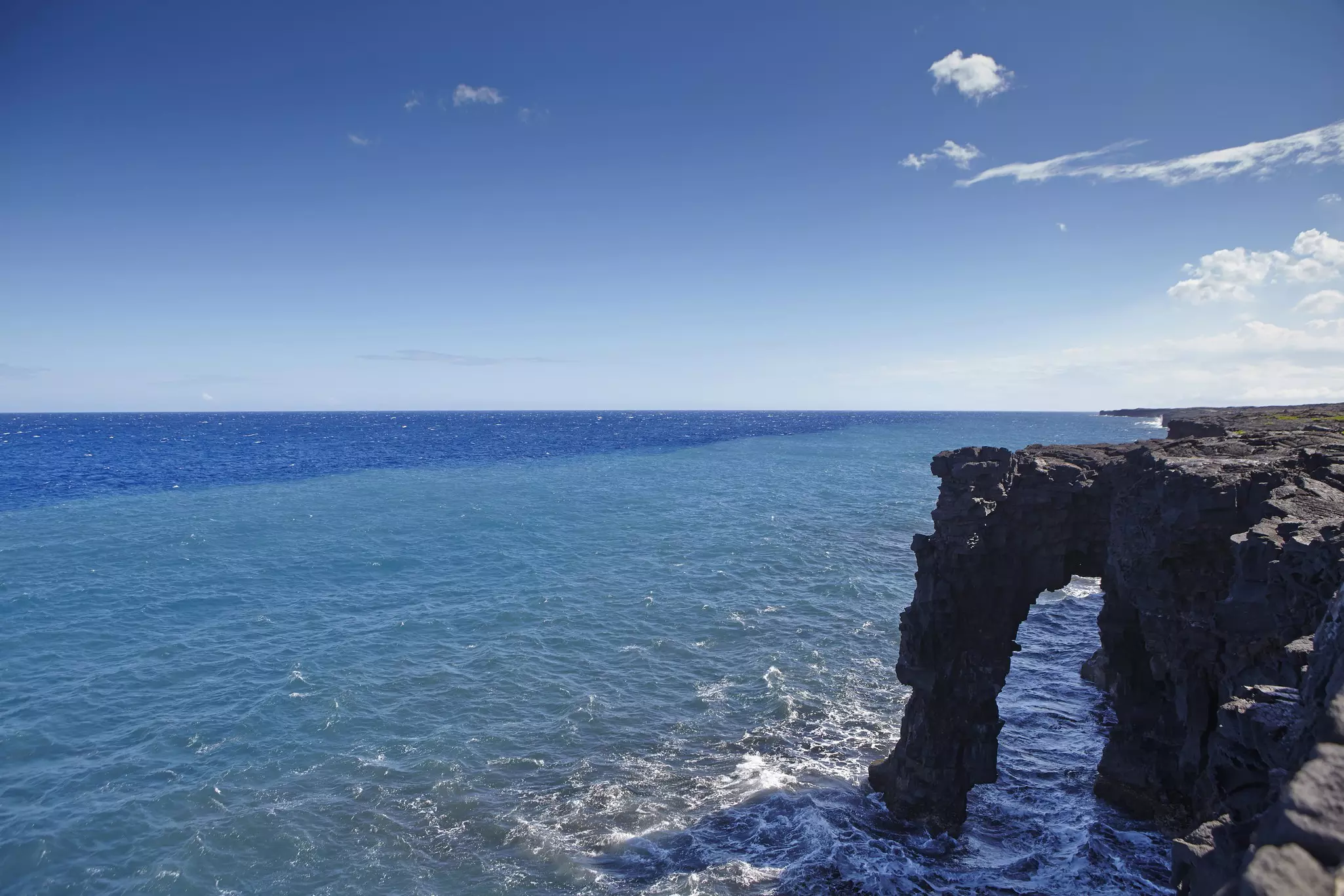 Rock arch and coastline with bright blue ocean to the left and into the distance on a sunny day.