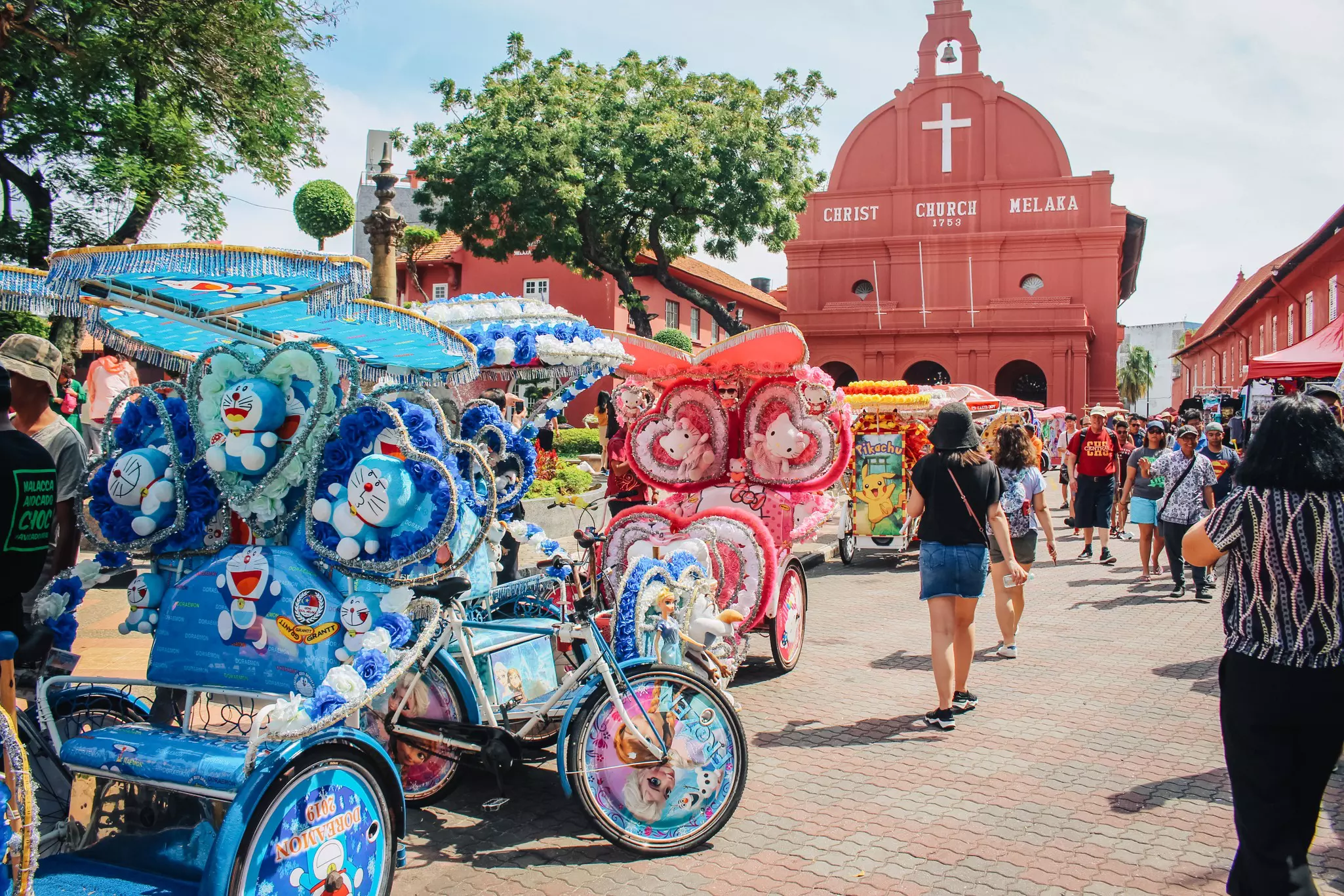 People in all different styles of clothing mill around in front of a church in an old town. Heavily decorated tri-shaw vehicles stand nearby ready to take passengers.