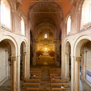 Elevated view down a stone nave with rounded arches and carved capitals toward a gilded altarpiece, a few people in pews
