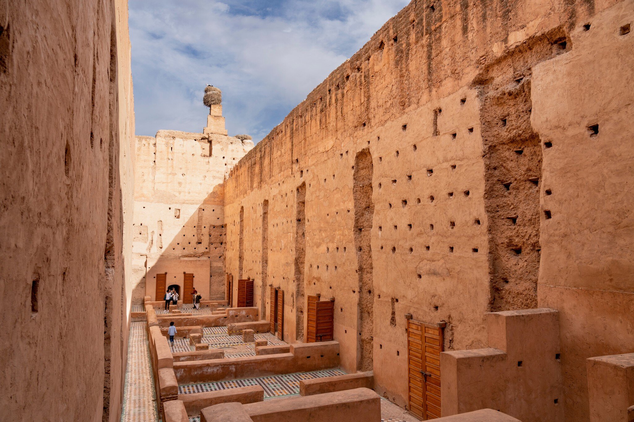 High ochre walls pocked with holes above tiled floor remnants and wooden doors, a large nest on a pillar top
