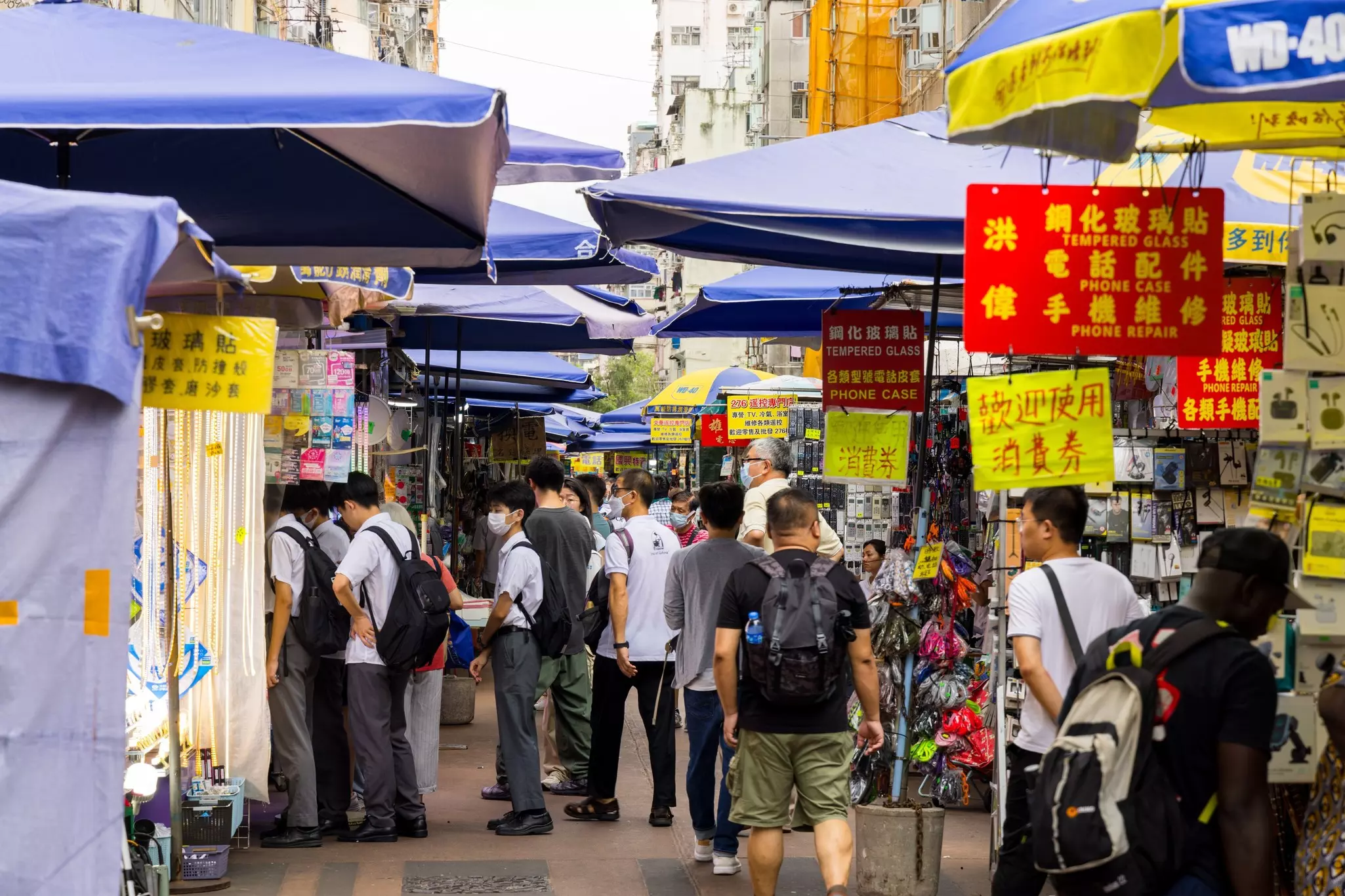 Busy alleyway with vendor stalls under blue umbreallas in Hong Kong.